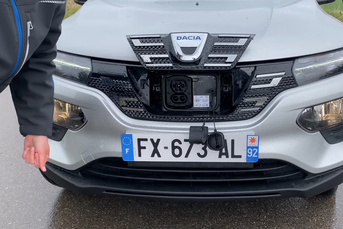 Une Dacia Spring bleue est branchée à une borne de recharge dans un village français. Des bâtiments en pierre et des fleurs colorées sont visibles en arrière-plan.
