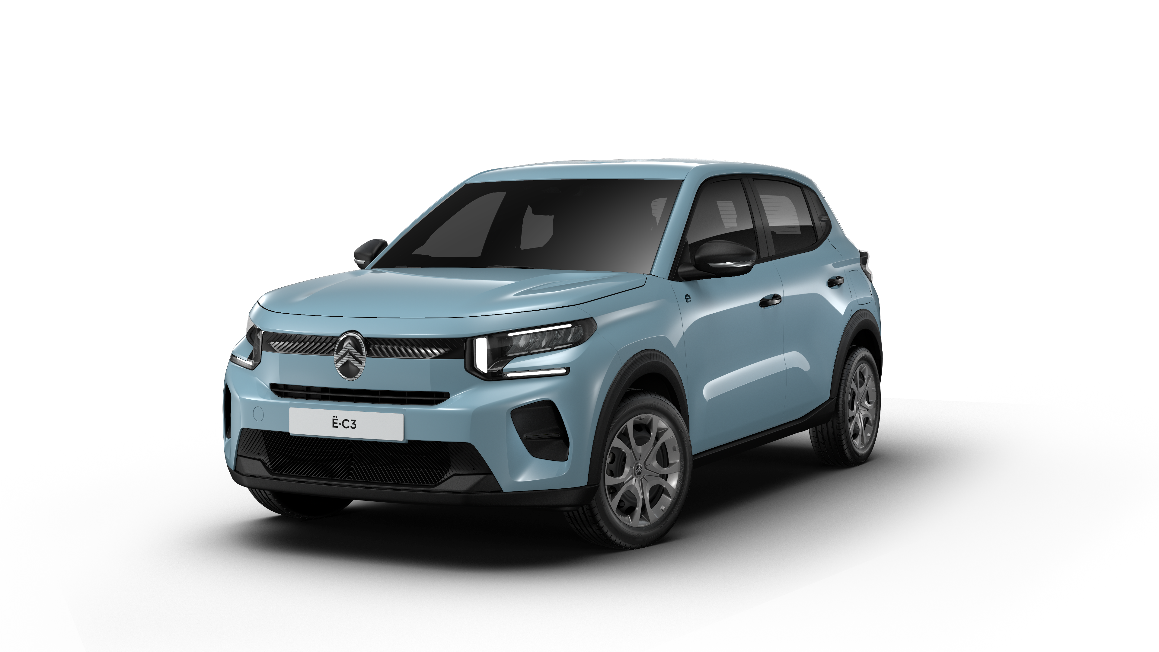 Une Citroën ë-C3 2026 de couleur pastel, en mouvement sur une route de banlieue, avec des maisons et des arbres en arrière-plan.