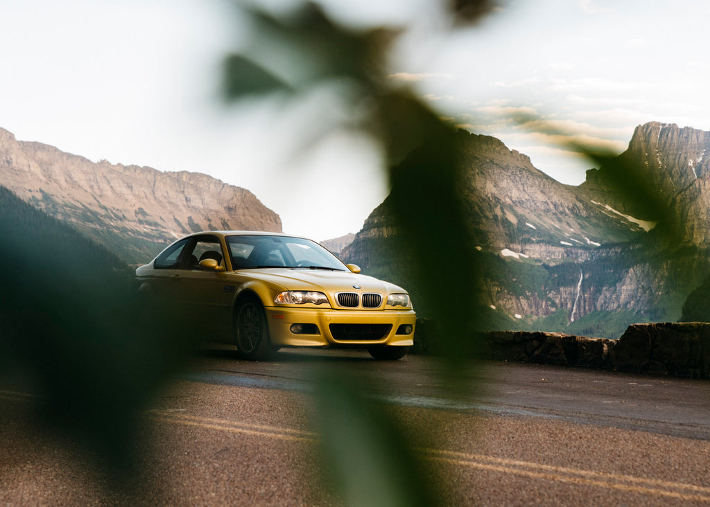 Une BMW M3 E46 argentée roule sur une route côtière sinueuse au coucher du soleil.