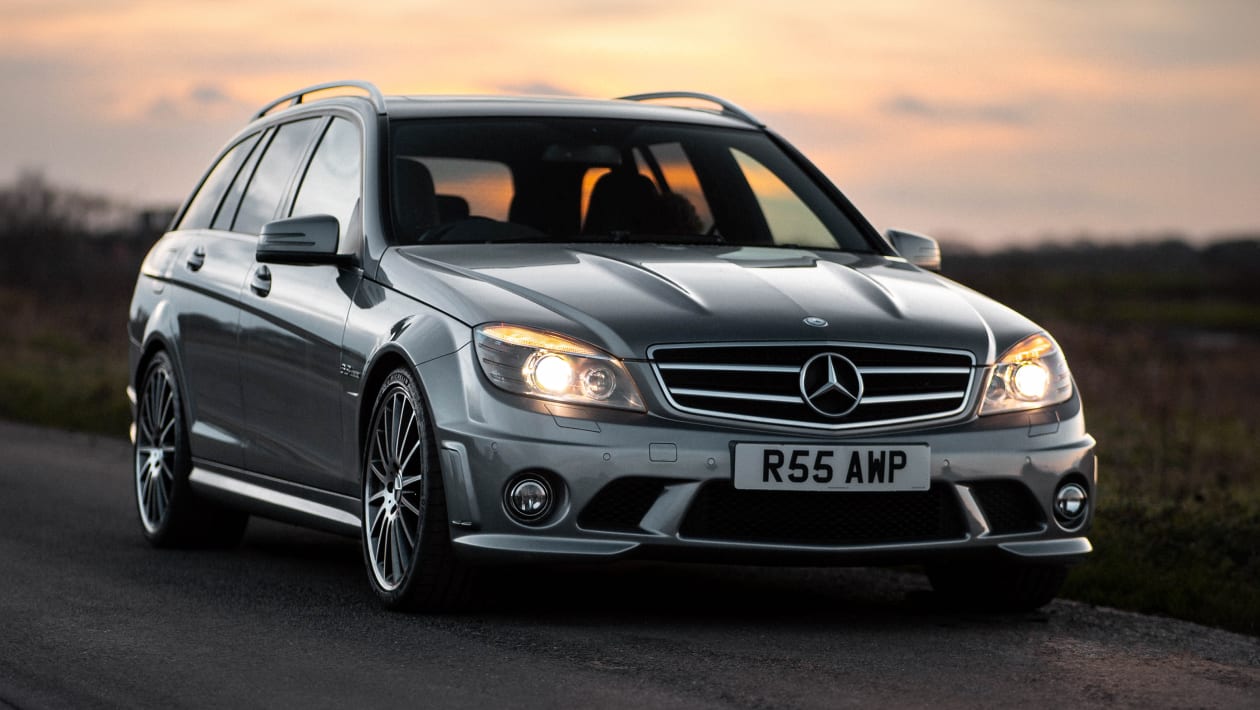 Une Mercedes C63 AMG W204 grise foncée en pleine accélération sur une route de campagne sinueuse, vue de face et légèrement de côté.