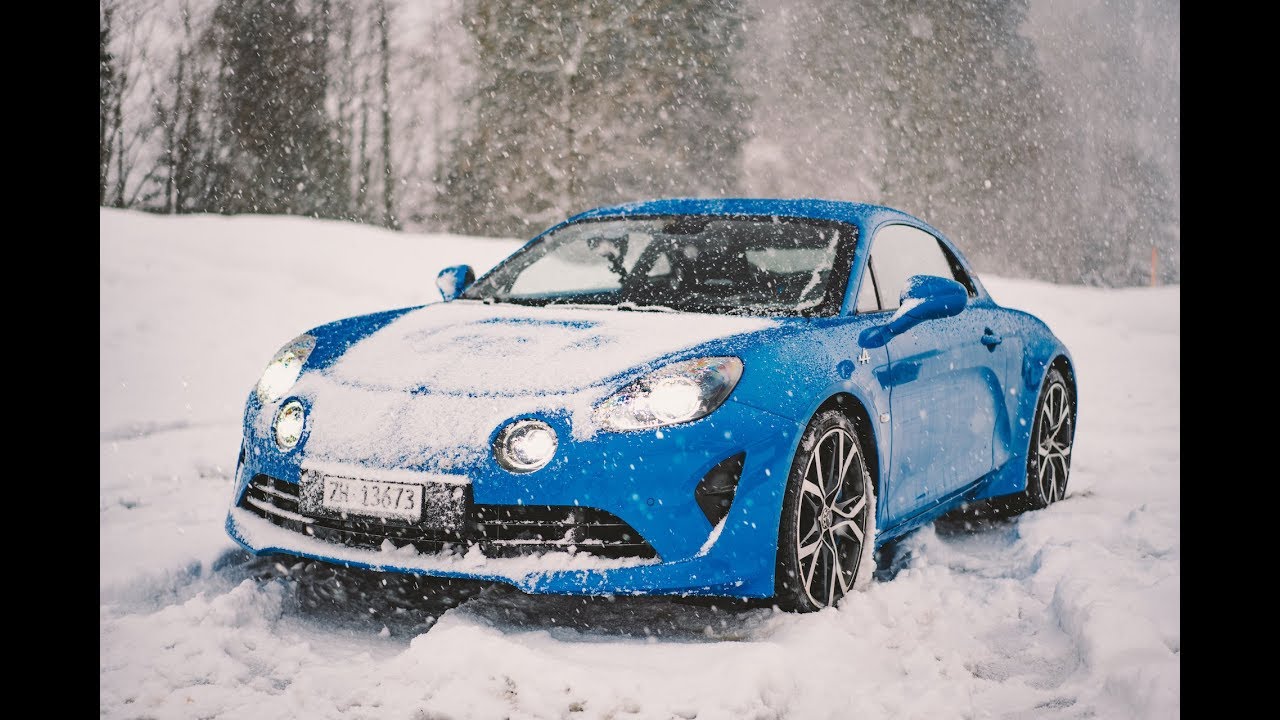 Une Alpine A110 bleue roulant sur une route couverte de neige, avec des arbres enneigés en arrière-plan.