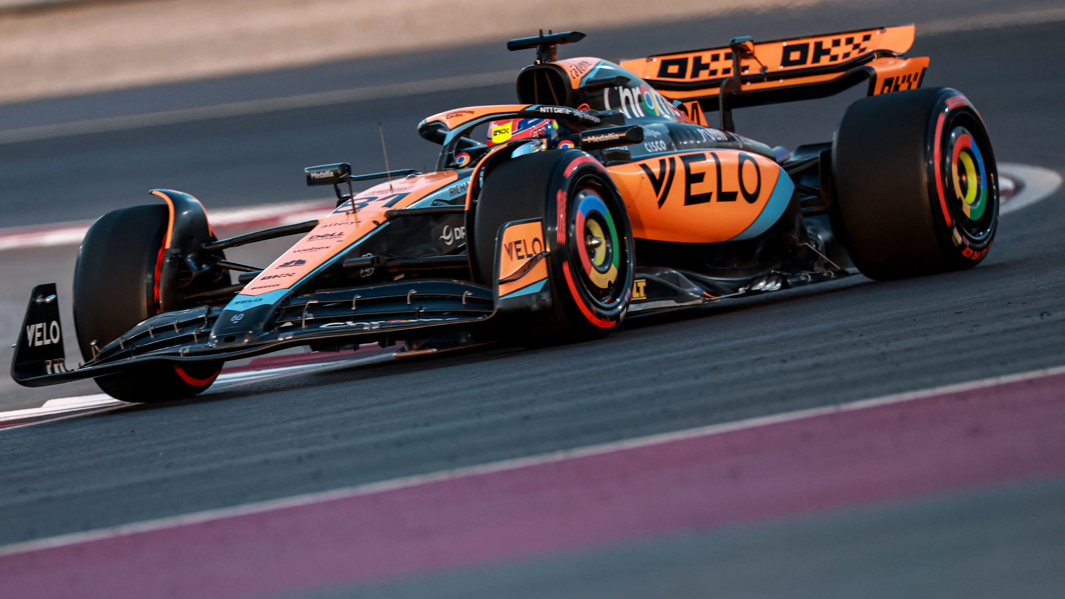 Une voiture de Formule 1 McLaren orange et noire, filant à toute vitesse sur une piste de course moderne sous un ciel clair.