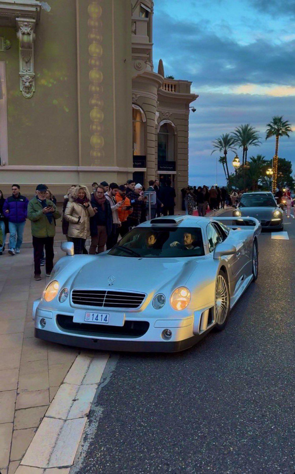 Une Mercedes-Benz CLK GTR garée devant un bâtiment luxueux à Monaco, avec Fernando Alonso posant à côté.