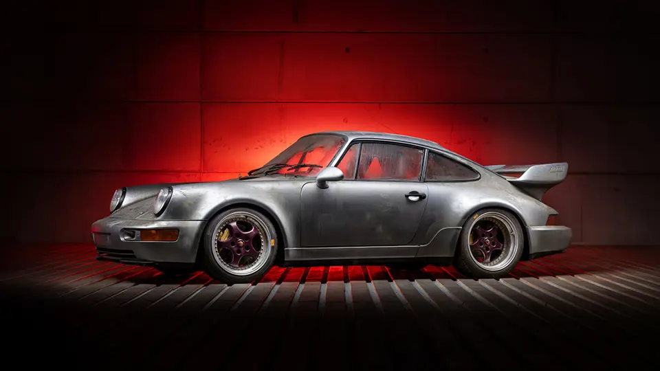 Une Porsche 911 Carrera RSR 3.8 version route rouge, avec son aileron arrière distinctif et ses passages de roues larges, garée sur une route sinueuse.