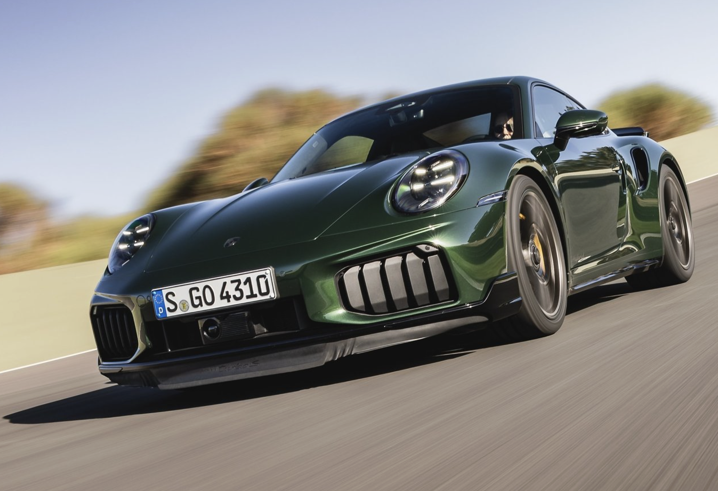Une Porsche 992 Turbo S de 2026, de couleur vive, roulant à grande vitesse sur une route de montagne sinueuse, avec un ciel dramatique en arrière-plan.