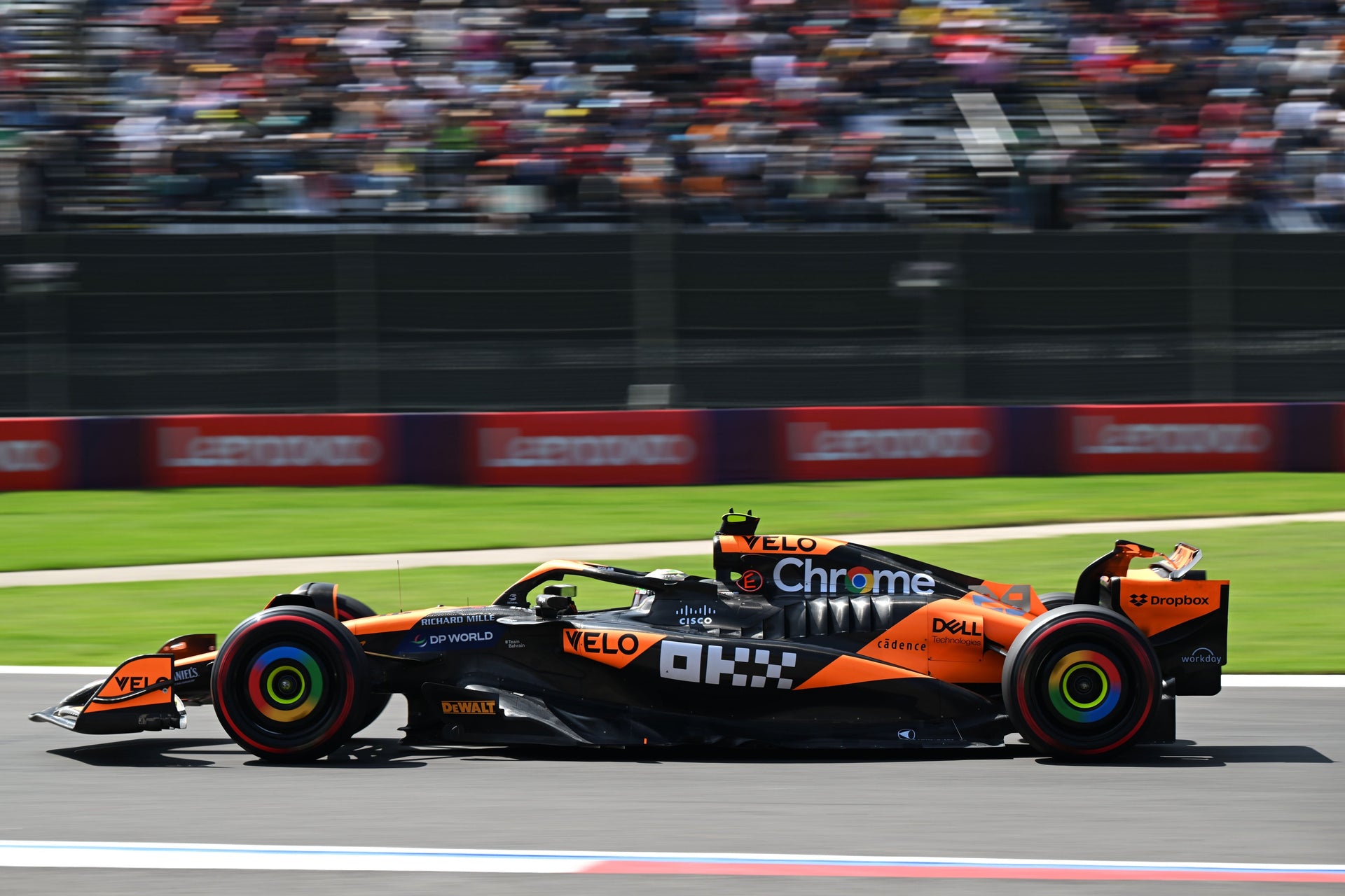 Une voiture de Formule 1 McLaren orange et noire en mouvement sur une piste de course, avec des tribunes en arrière-plan.