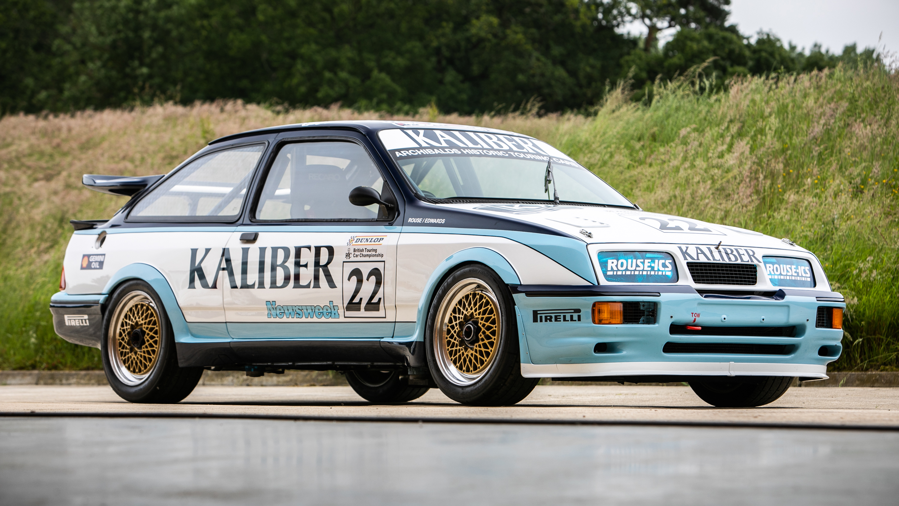 Une Ford Sierra RS500 Cosworth blanche avec des bandes bleues et rouges, en train de courir sur une piste, avec d'autres voitures de course en arrière-plan.