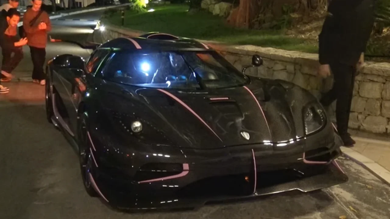 Une Koenigsegg One:1 rouge et noire roulant dans les rues de Monaco, avec des voitures de police en arrière-plan, suggérant une scène de poursuite.
