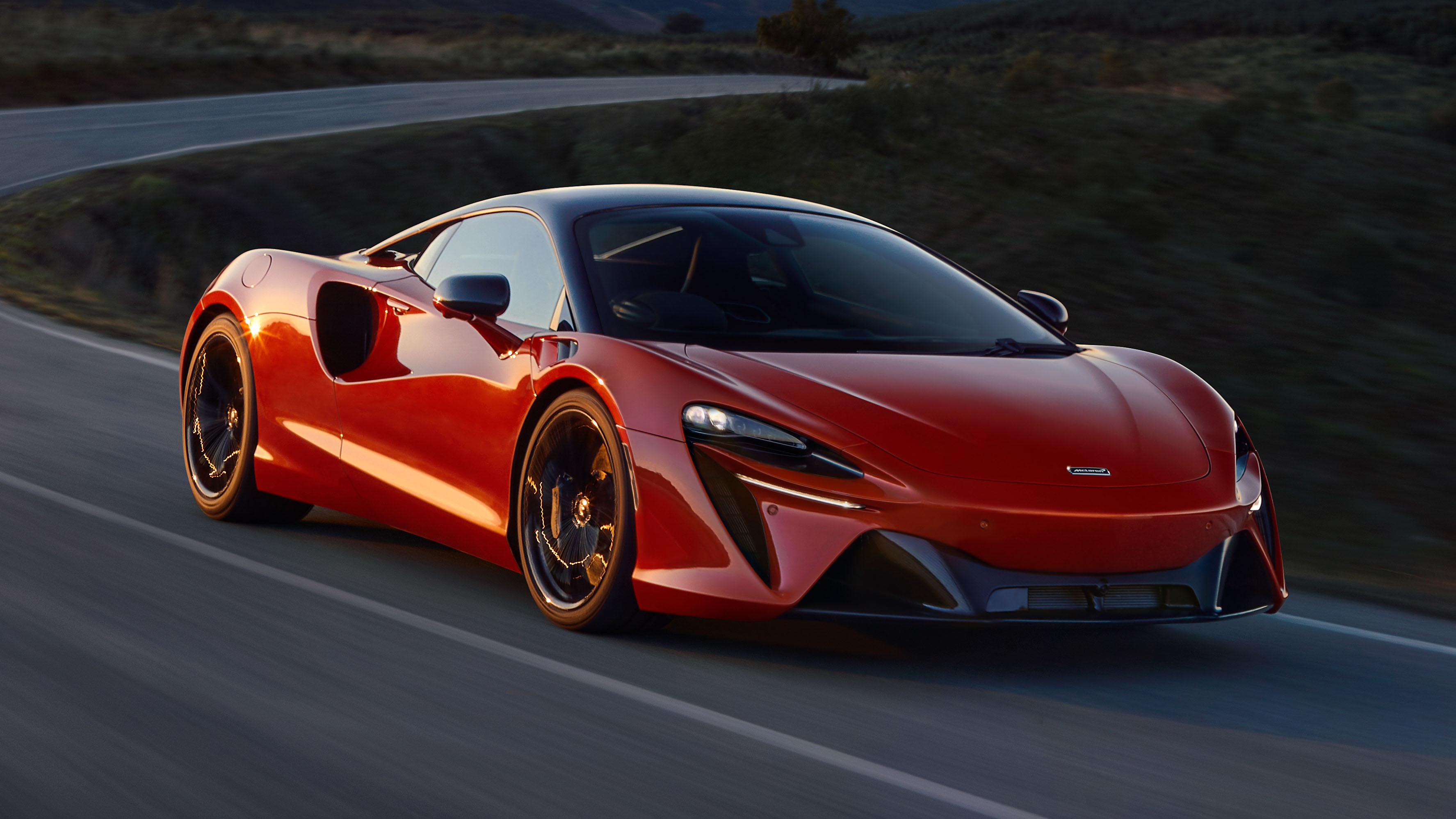 Une McLaren Artura rouge vif en mouvement sur une route sinueuse, son design élégant et sportif mis en valeur par un arrière-plan flou.
