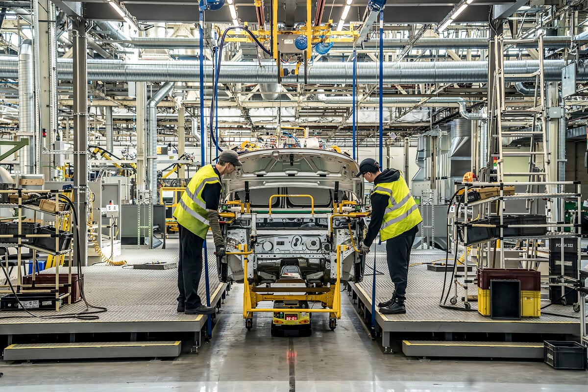 Vue aérienne d'une usine de fabrication automobile moderne et vaste, avec des toits gris et des panneaux solaires, située dans un paysage industriel verdoyant.