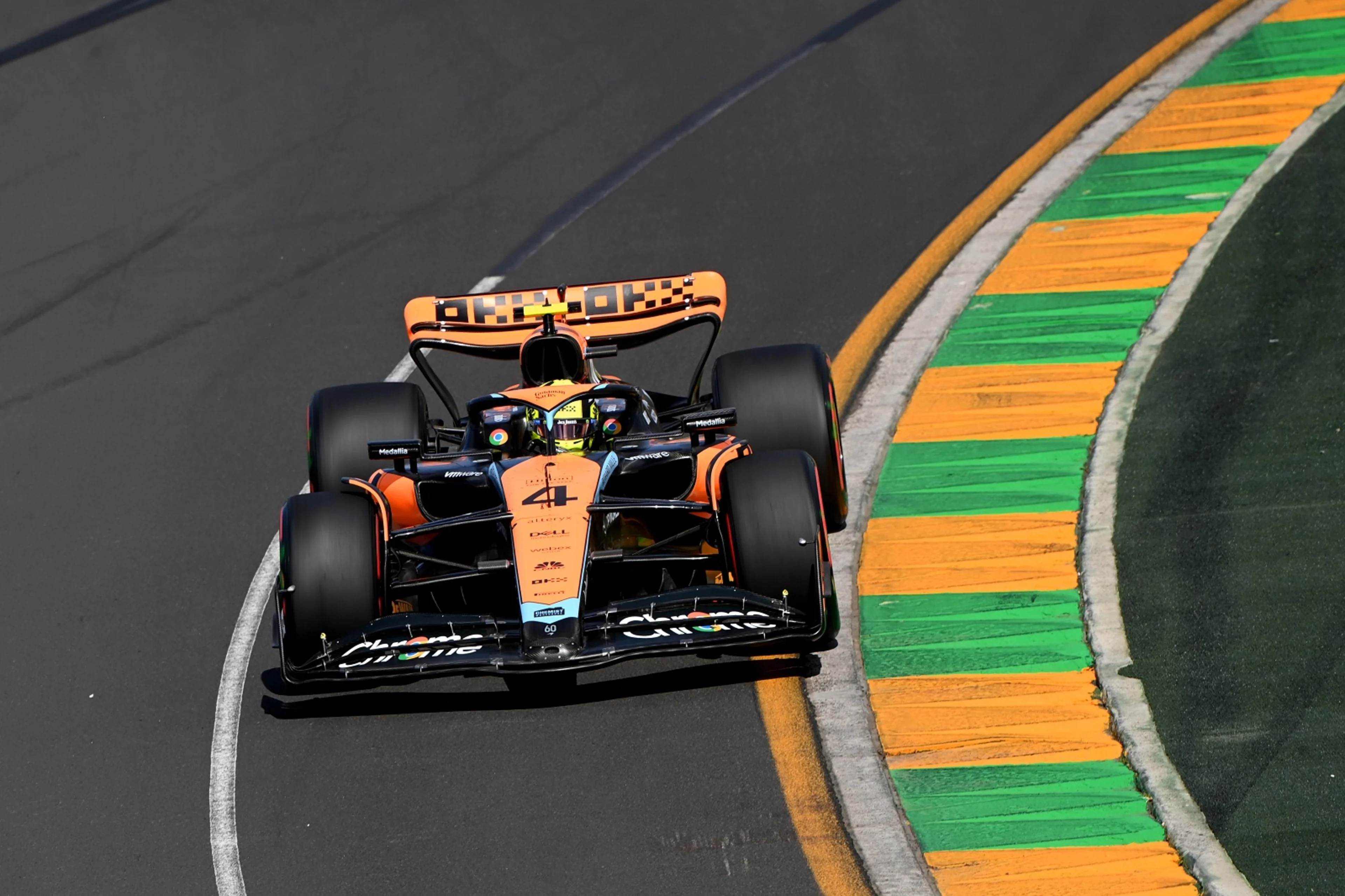 Une voiture de Formule 1 McLaren orange et noire sur une piste de course, avec des mécaniciens et des ingénieurs observant depuis les stands.