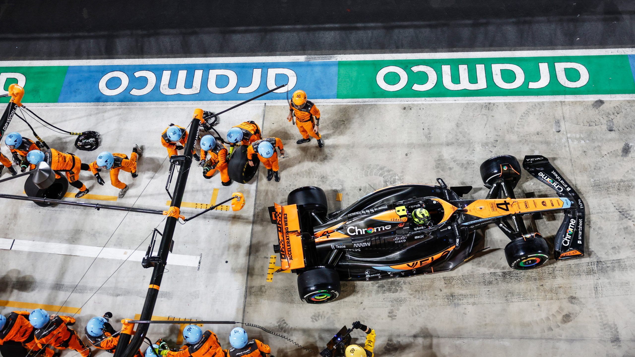Des mécaniciens de l'équipe McLaren en uniforme orange et noir, changeant rapidement les pneus d'une voiture de Formule 1 dans la voie des stands.