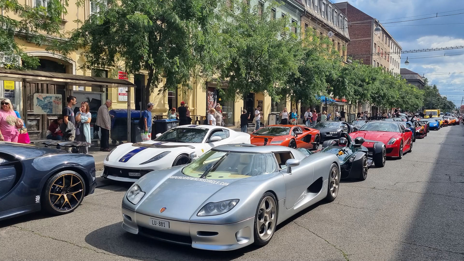 Vue aérienne d'un événement public avec un convoi d'hypercars, montrant des barrières, du personnel de sécurité et des policiers gérant la foule.