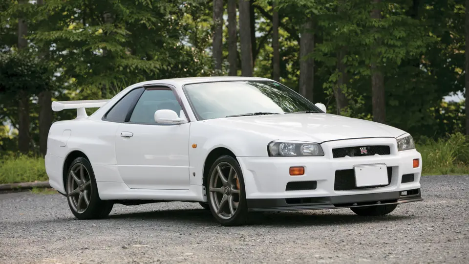Une Nissan Skyline GT-R R34 V-Spec II Nür bleue, exposée sous les projecteurs lors d'une vente aux enchères, avec des spectateurs en arrière-plan.