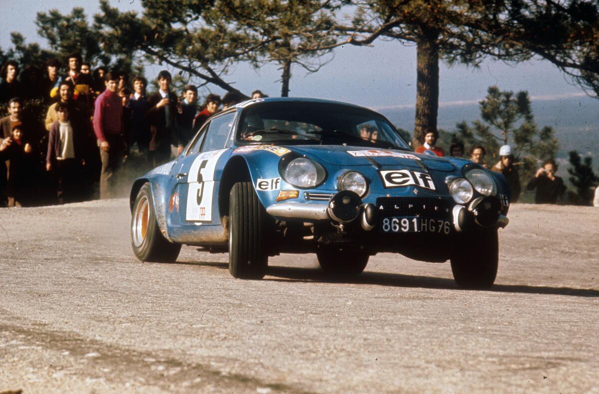 Patrick Tambay, un homme souriant, assis dans une Alpine A110 1600 S bleue, interagissant avec des fans lors du Tour Auto Optic 2000.