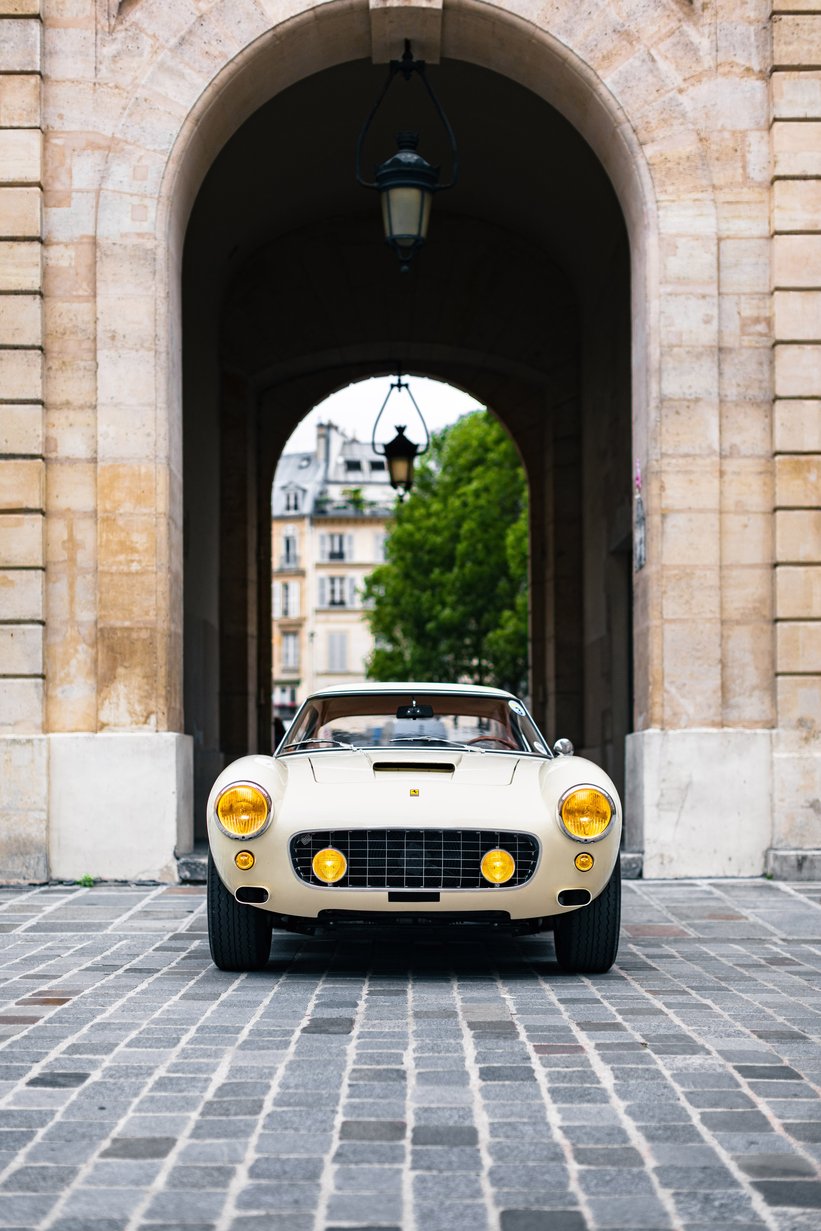 Une Ferrari 250 GT SWB rouge de 1960 en mouvement sur une route de campagne, avec des spectateurs en arrière-plan, lors du Tour Auto Optic 2000.