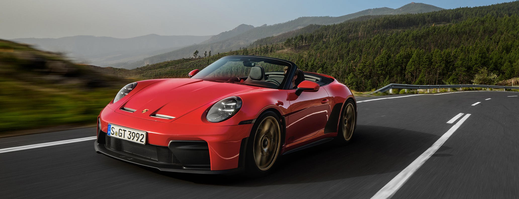 Une Porsche 911 GT3 S/C cabriolet rouge vif roulant sur une route côtière au coucher du soleil, avec l'océan en arrière-plan.