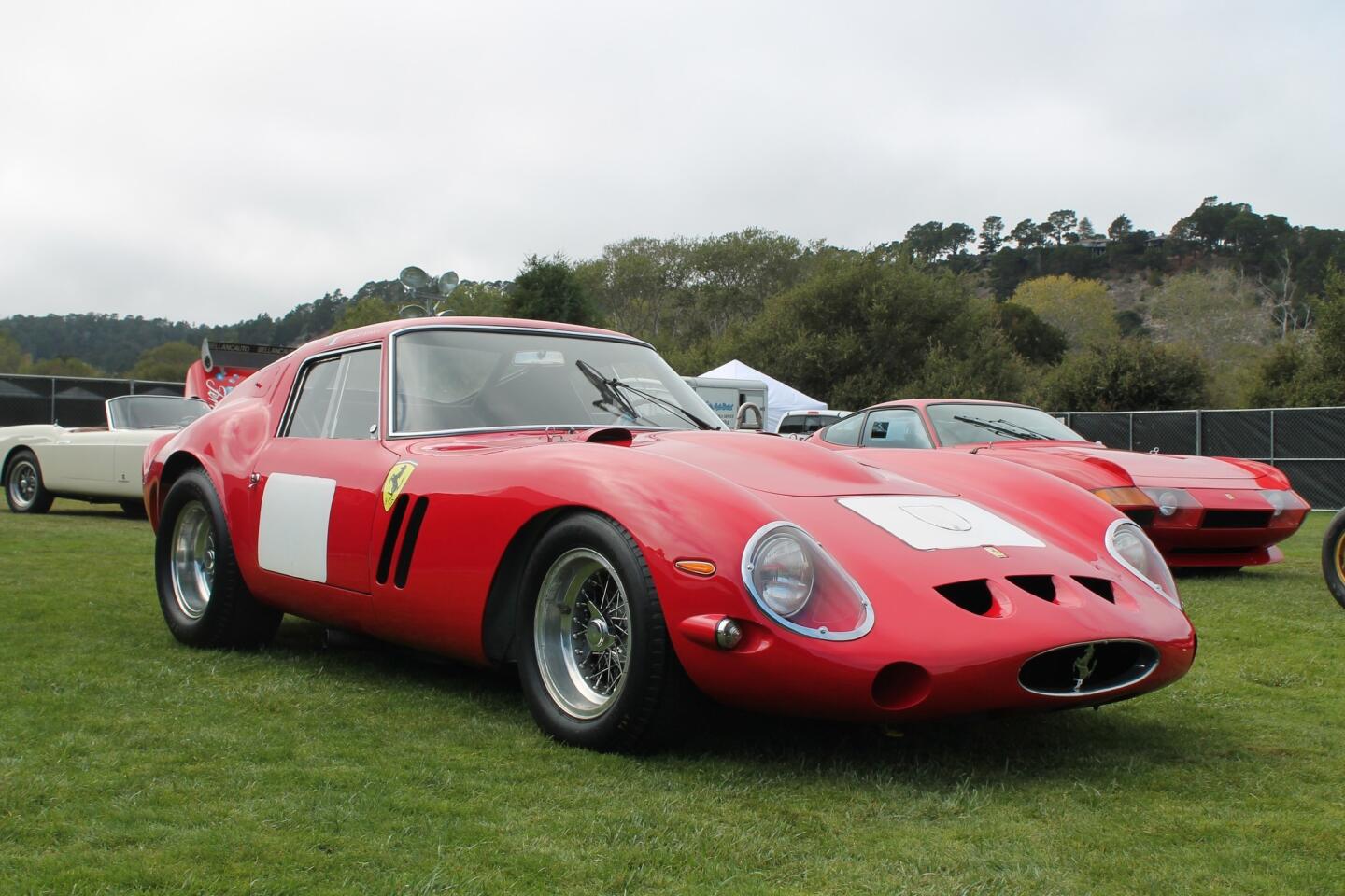 Gros plan d'une Ferrari 250 GTO rouge brillante sur un podium d'enchères, avec des collectionneurs flous en arrière-plan, lors de la Monterey Car Week.