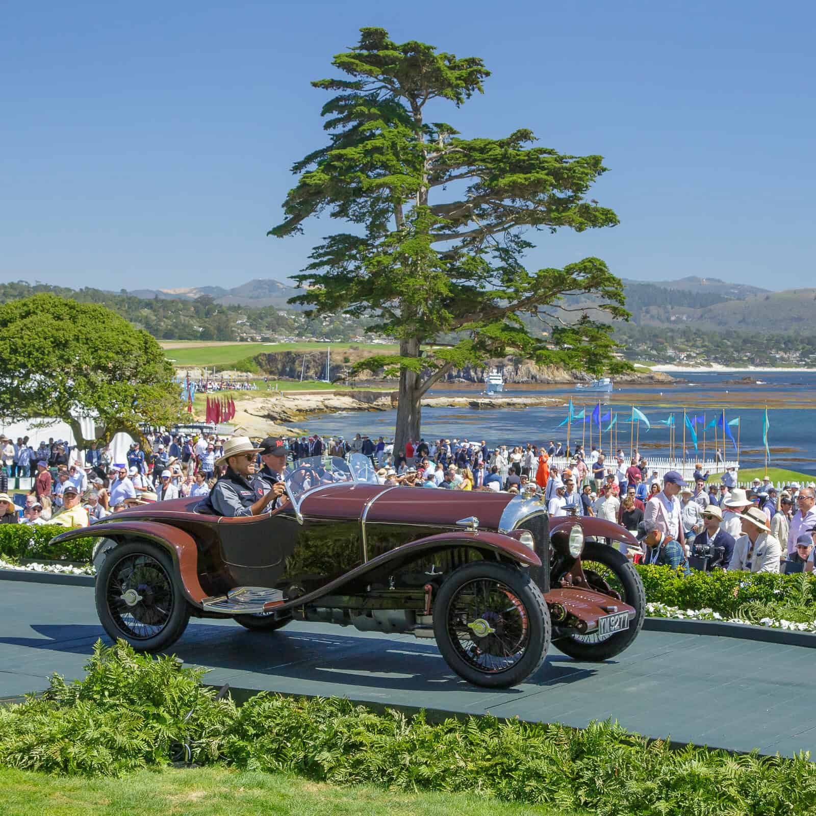 Vue aérienne du Concours d'Élégance de Pebble Beach, montrant des supercars classiques impeccablement alignées sur une pelouse verte, avec l'océan Pacifique en arrière-plan.