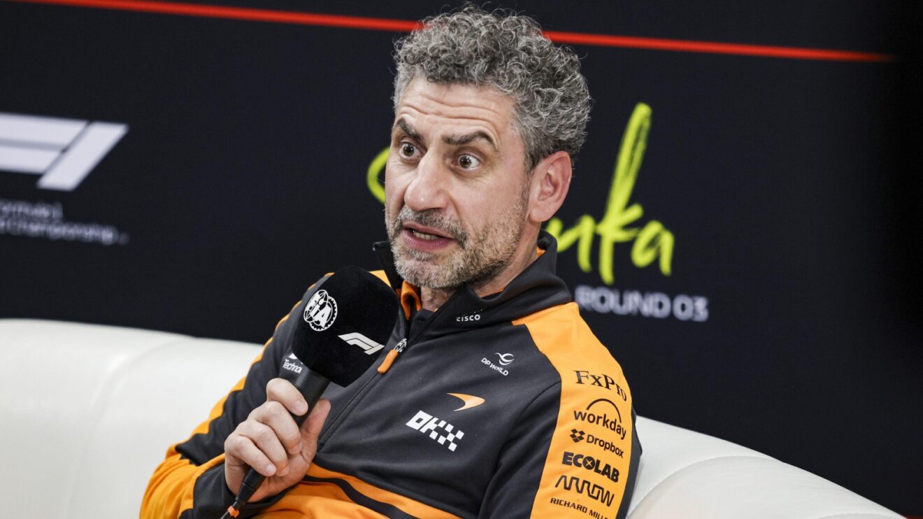 Andrea Stella, directeur de McLaren, en pleine réflexion dans le paddock, avec des monoplaces de Formule 1 en arrière-plan.