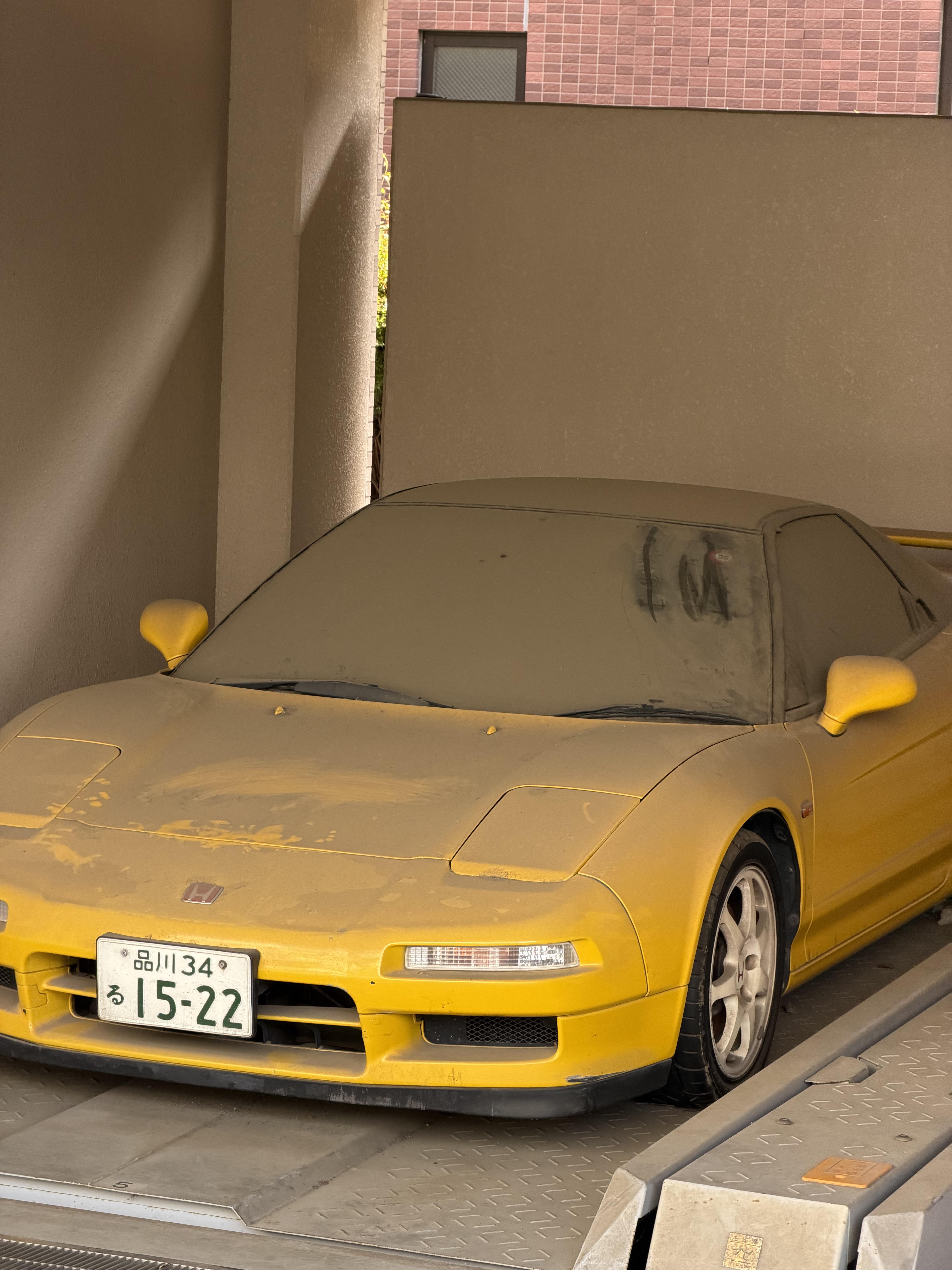 Une Honda NSX-R jaune abandonnée et poussiéreuse, garée dans un parking résidentiel à Tokyo.
