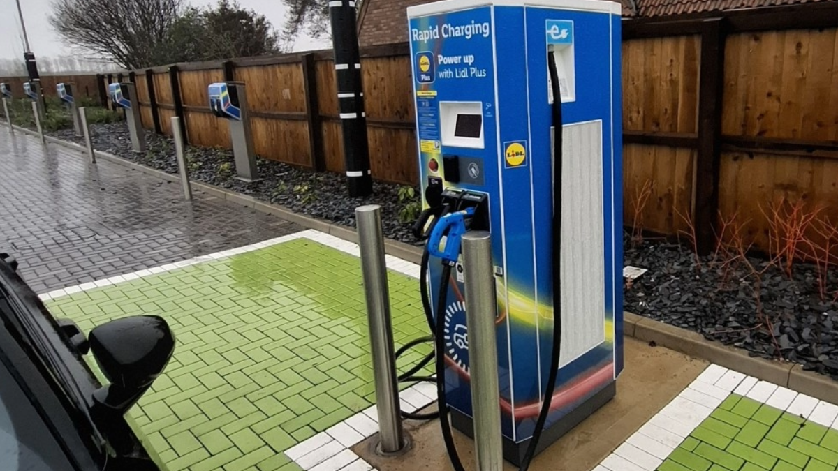 Une voiture électrique en charge sur une borne moderne et propre, située sur un parking de supermarché Lidl, avec le bâtiment du magasin en arrière-plan.