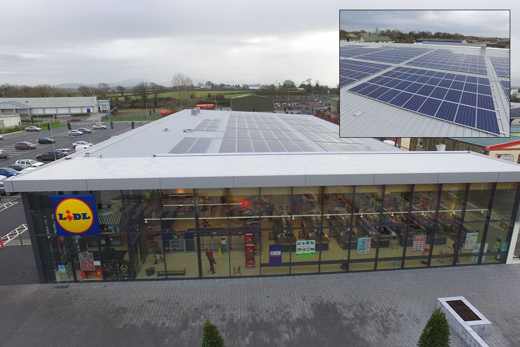 Vue aérienne d'un supermarché Lidl avec de nombreux panneaux solaires brillants sur son toit plat, sous un ciel bleu clair.