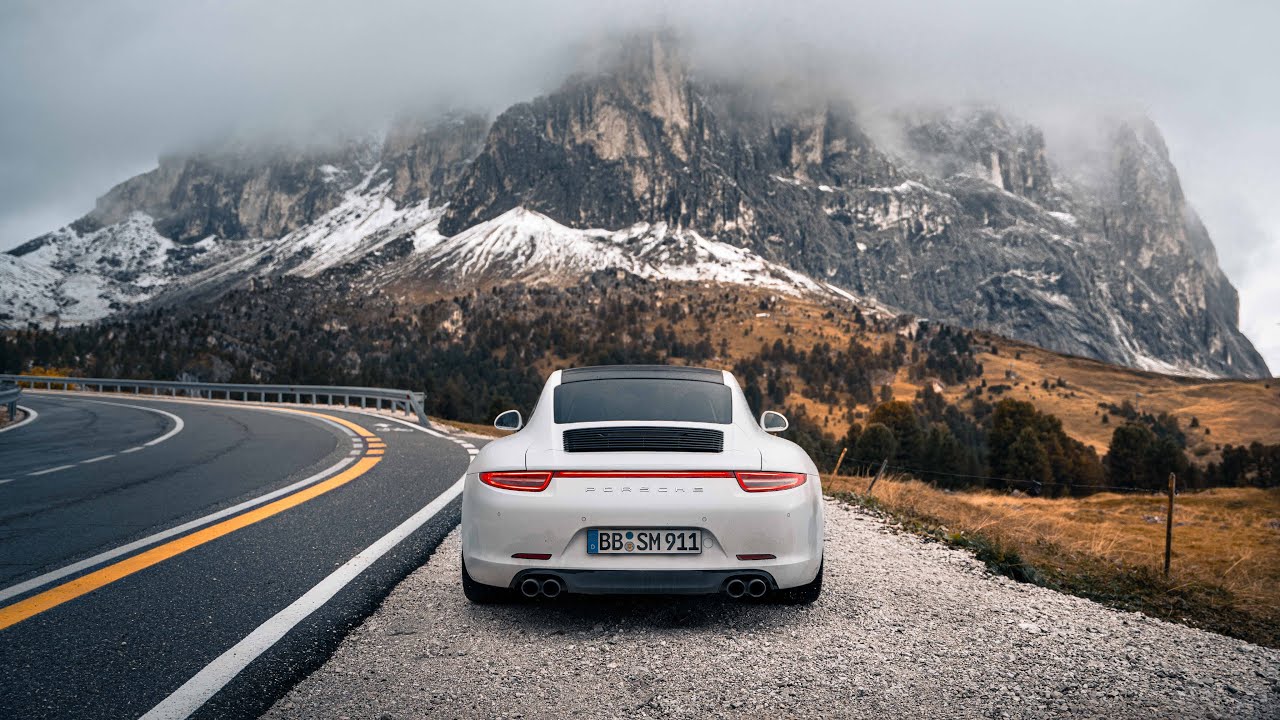 Une Porsche 911 rouge roulant sur une route côtière au coucher du soleil, symbolisant la résilience des modèles thermiques de la marque.