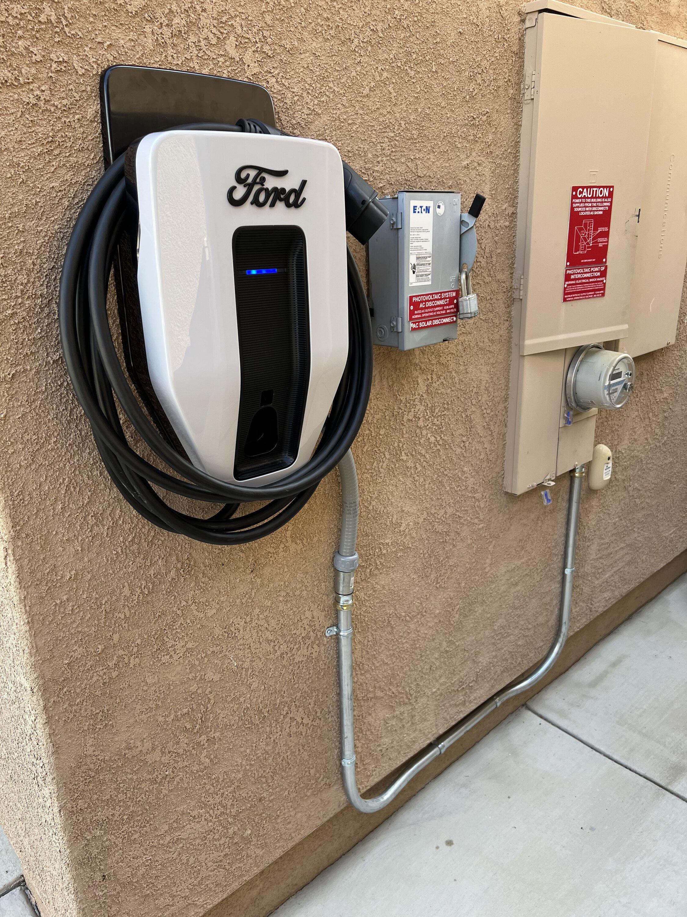 Une borne de recharge Ford Charge Station Pro blanche et noire, fixée au mur d'un garage moderne et lumineux, avec un câble branché à une Ford Mustang Mach-E garée à proximité.