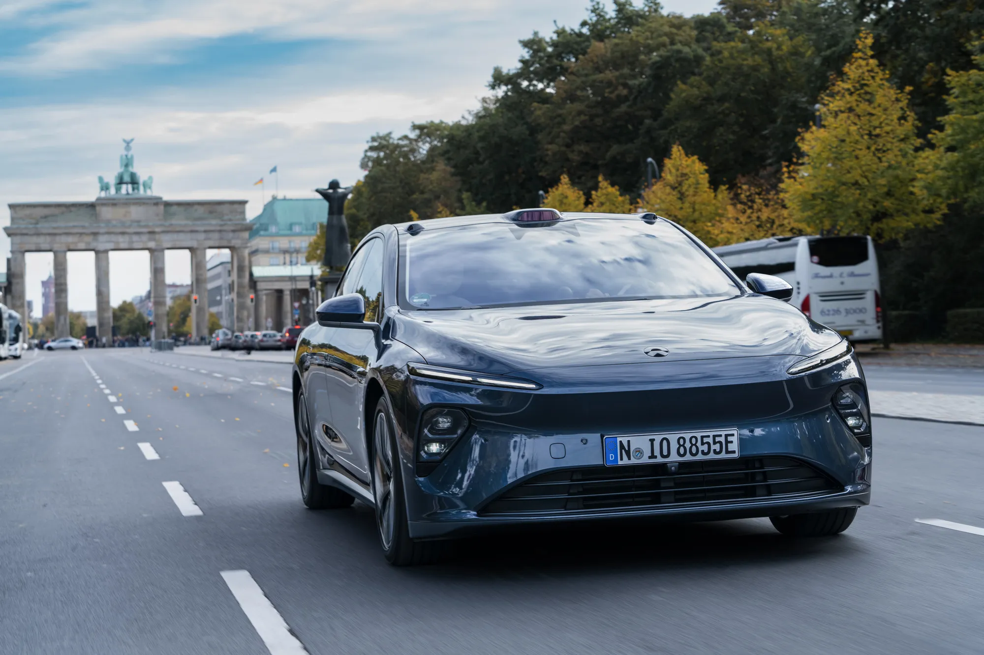 Une voiture électrique Nio de grande taille circulant dans une rue étroite et historique d'une ville européenne, avec des bâtiments anciens et des pavés. La voiture semble un peu grande pour l'environnement.
