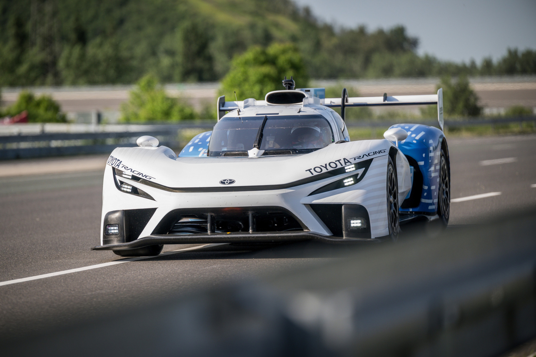 Une voiture de course futuriste, le prototype Toyota TR LH2 Racing, roule sur une piste mouillée au Mans, avec des tribunes et des spectateurs flous en arrière-plan.