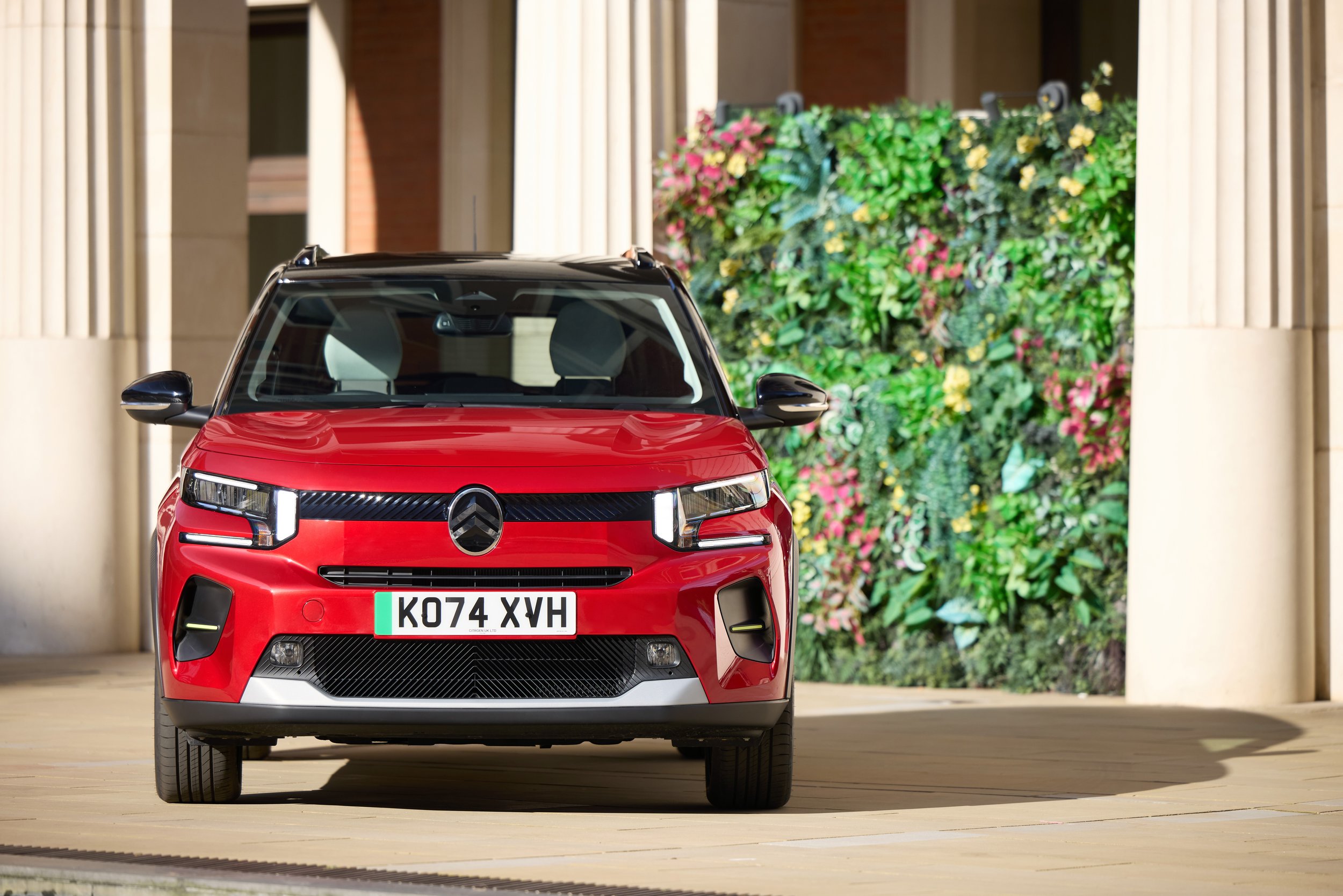 Une Citroën ë-C3 blanche roulant sur une route de ville, avec des bâtiments modernes en arrière-plan.