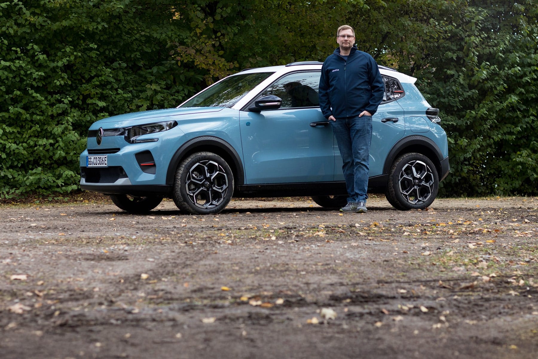 Une Citroën ë-C3 électrique rouge vif roulant sur une route de campagne sinueuse, avec des champs et des fermes en arrière-plan.