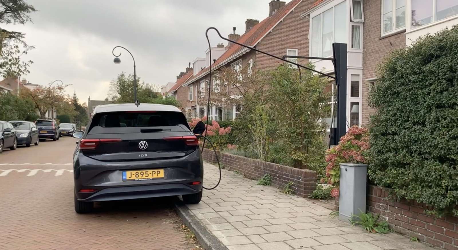 Un câble de recharge orange pour véhicule électrique est posé sur un trottoir, recouvert d'une protection jaune pour éviter les chutes. En arrière-plan flou, une voiture électrique est garée devant un bâtiment français.