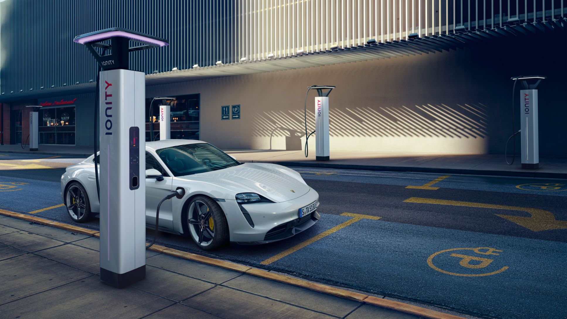 Une Porsche Taycan noire en charge sur une borne électrique, avec des lumières de ville en arrière-plan.