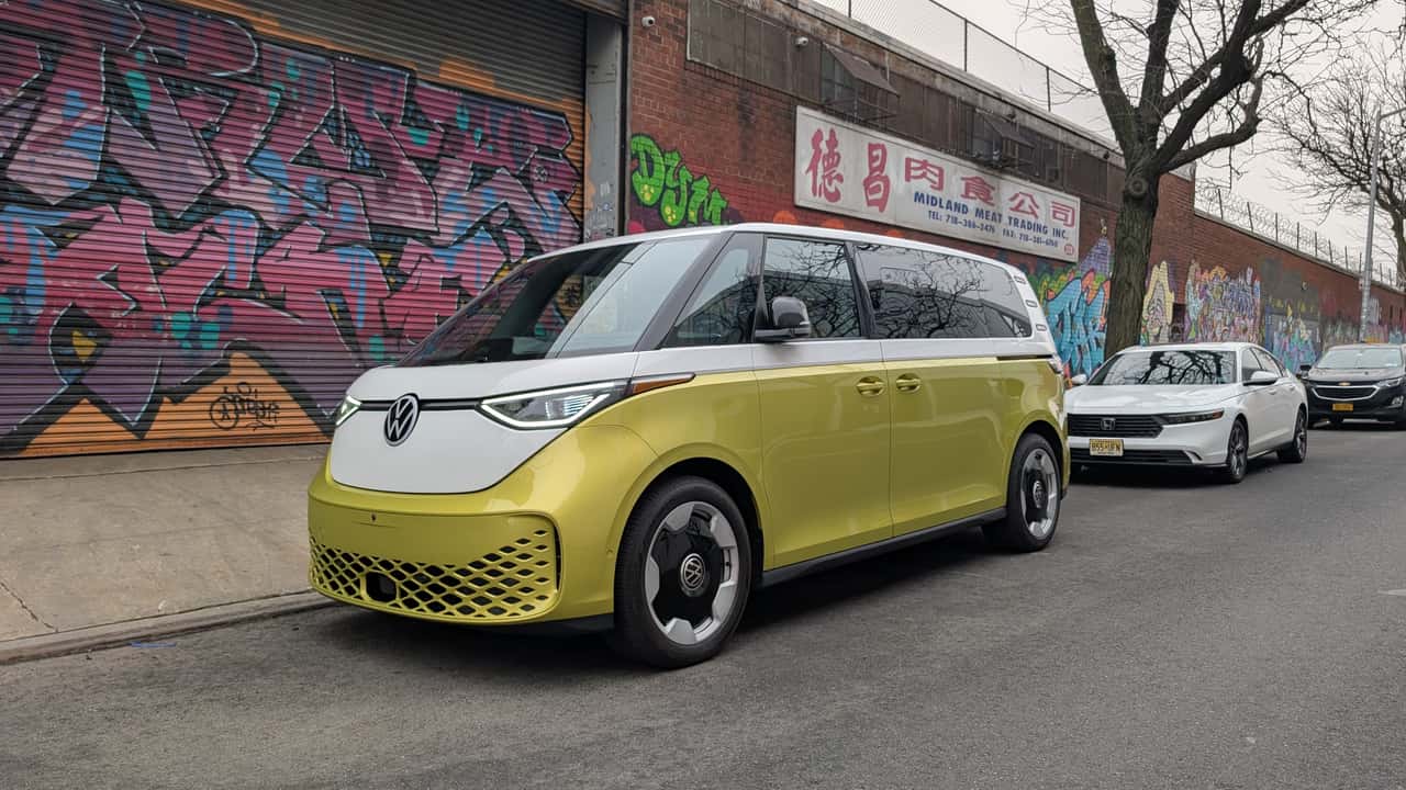 Un Volkswagen ID.Buzz blanc et jaune garé dans une rue de ville animée, avec des bâtiments modernes en arrière-plan.