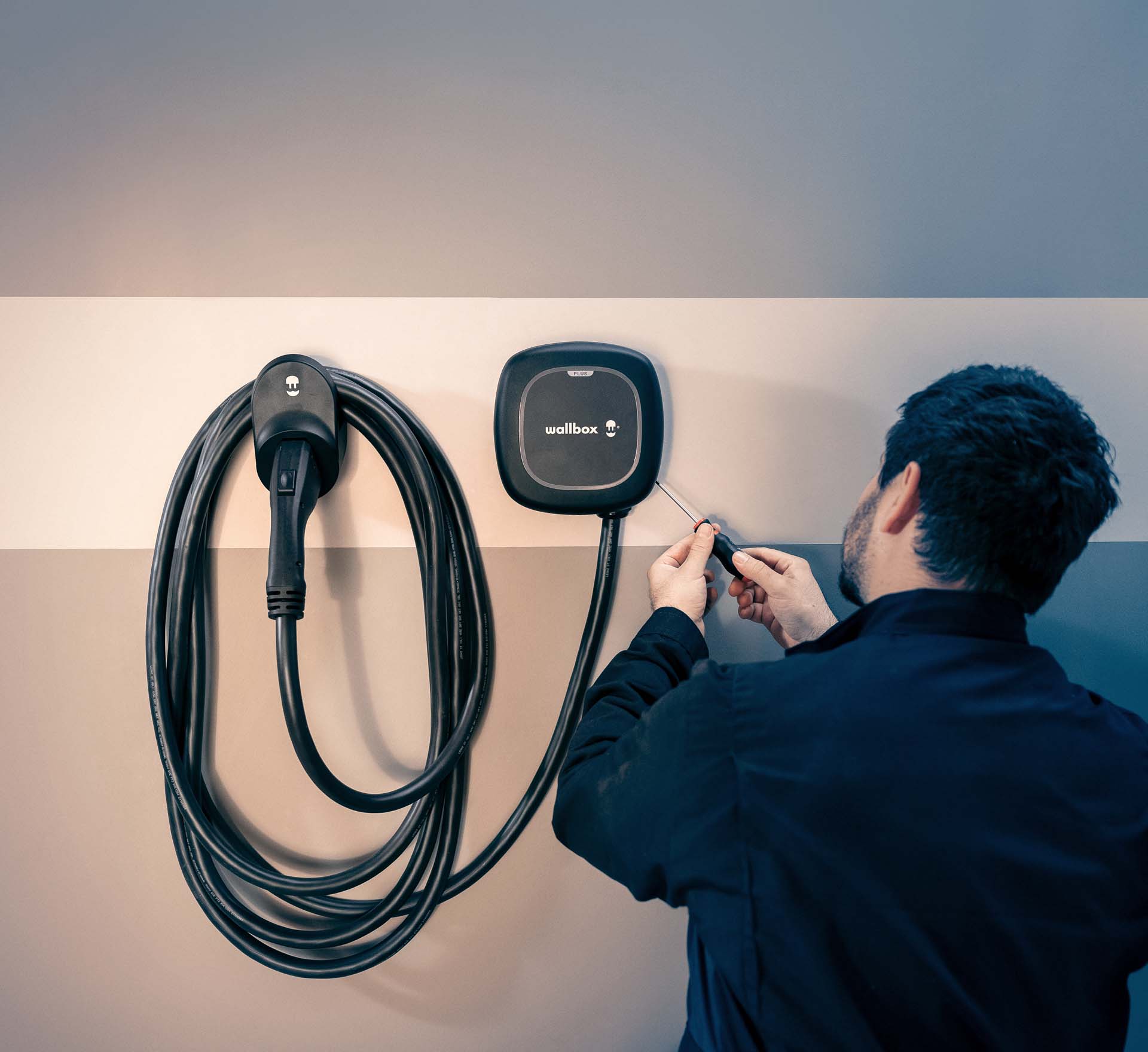 Photographie détaillée d'une borne de recharge murale (wallbox) installée dans un garage, avec un câble de recharge en cours de manipulation.