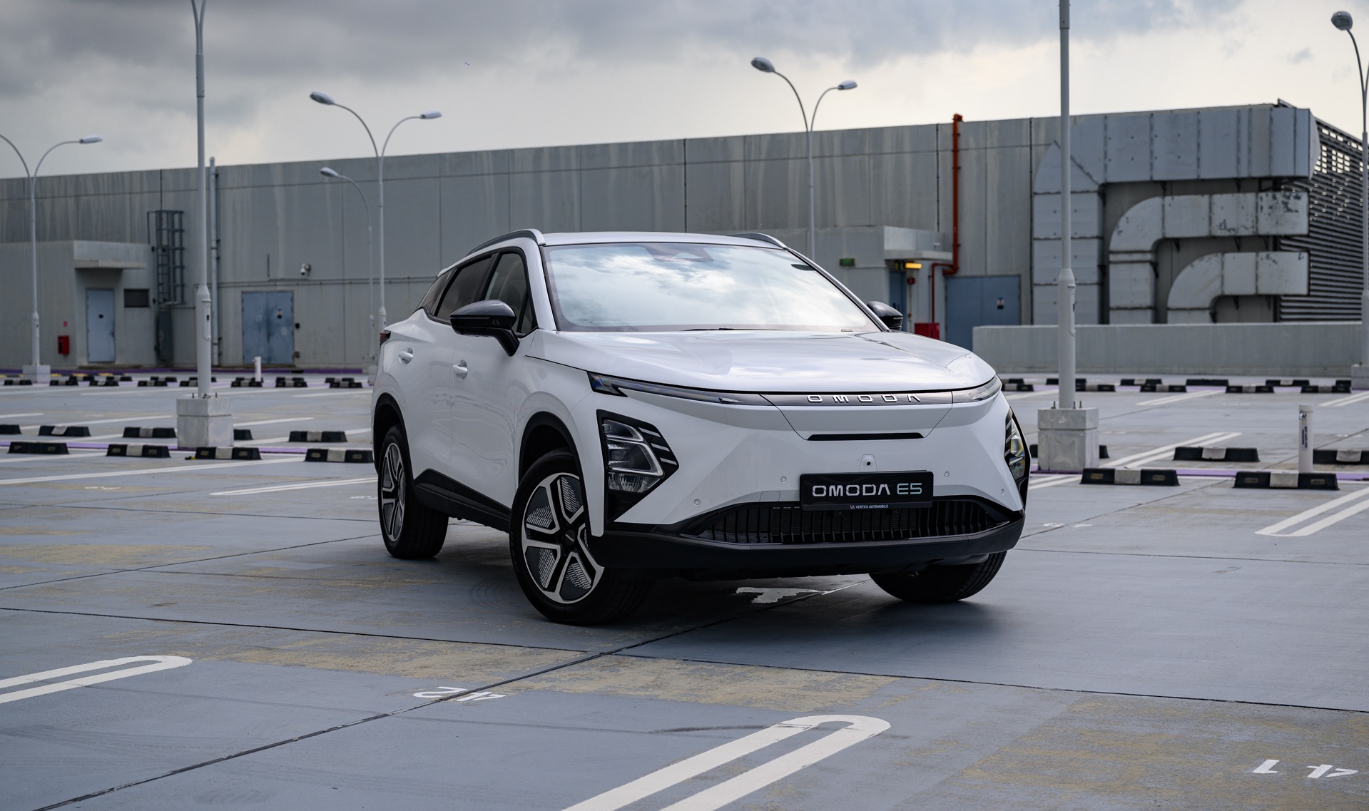Une voiture électrique Chery Omoda E5 blanche et élégante roule sur une route de ville moderne au Canada, avec des bâtiments contemporains en arrière-plan.