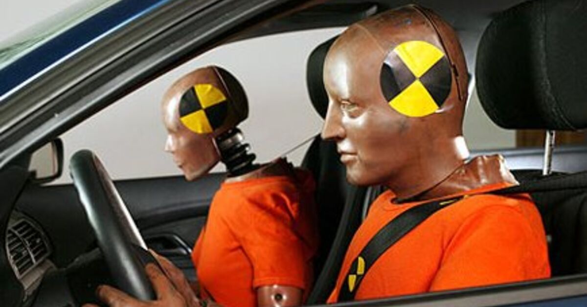 Deux mannequins de crash test, un adulte et un enfant, sont assis dans l'habitacle d'une voiture, prêts pour un test d'impact frontal. Des capteurs et des fils sont visibles sur les mannequins.