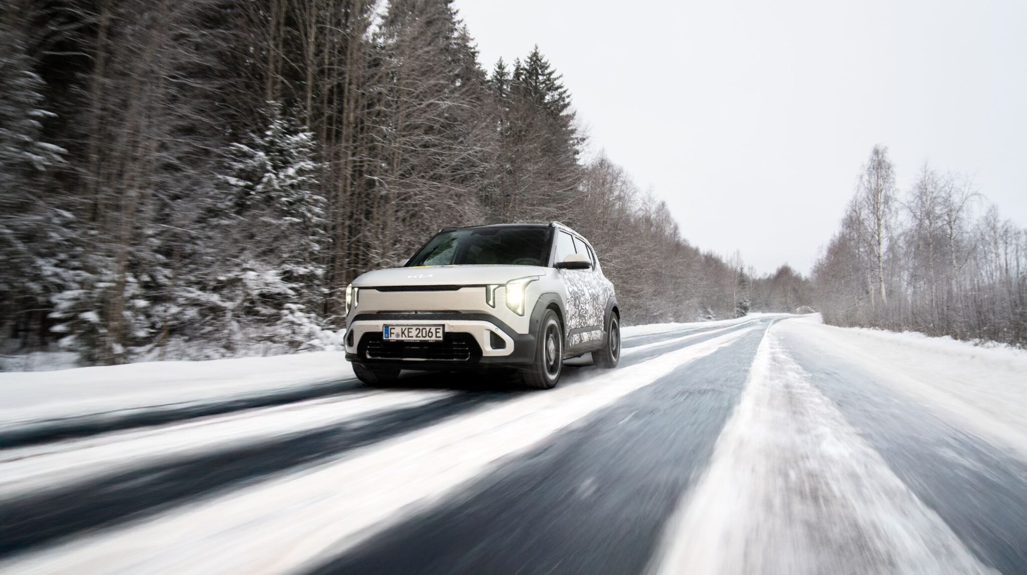 Un Kia EV2 blanc roulant sur une route enneigée et glacée dans un paysage montagneux norvégien en hiver.