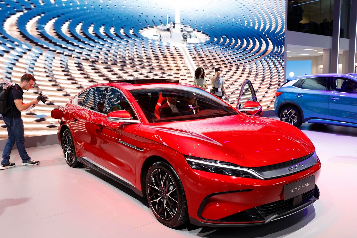 Une voiture électrique chinoise moderne, comme une MG4, circulant dans une rue de ville européenne avec des bâtiments historiques en arrière-plan.