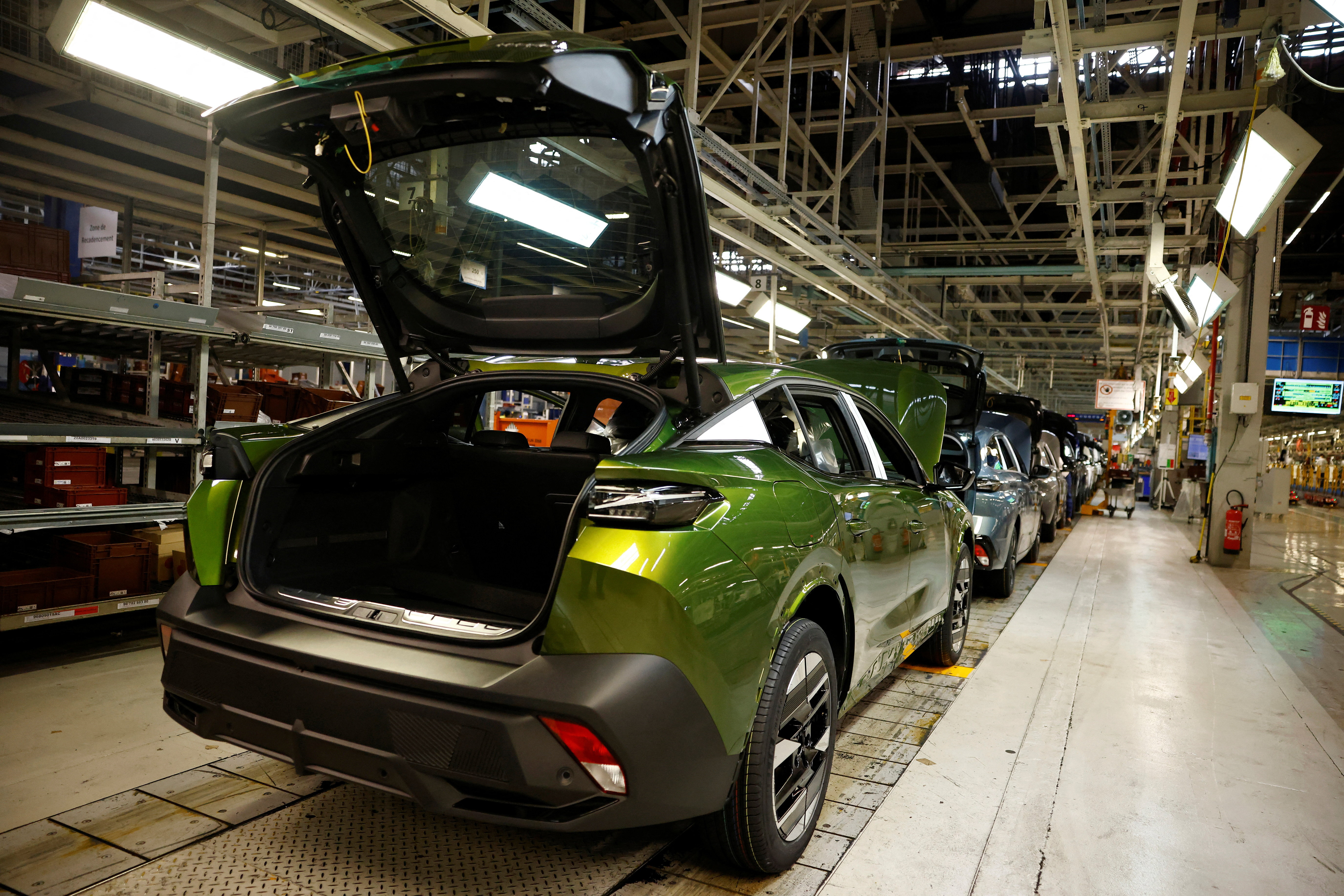 Une vue aérienne d'une usine Stellantis moderne et lumineuse à Mulhouse, montrant des bras robotiques assemblant des châssis de voitures électriques sur une ligne de production automatisée.