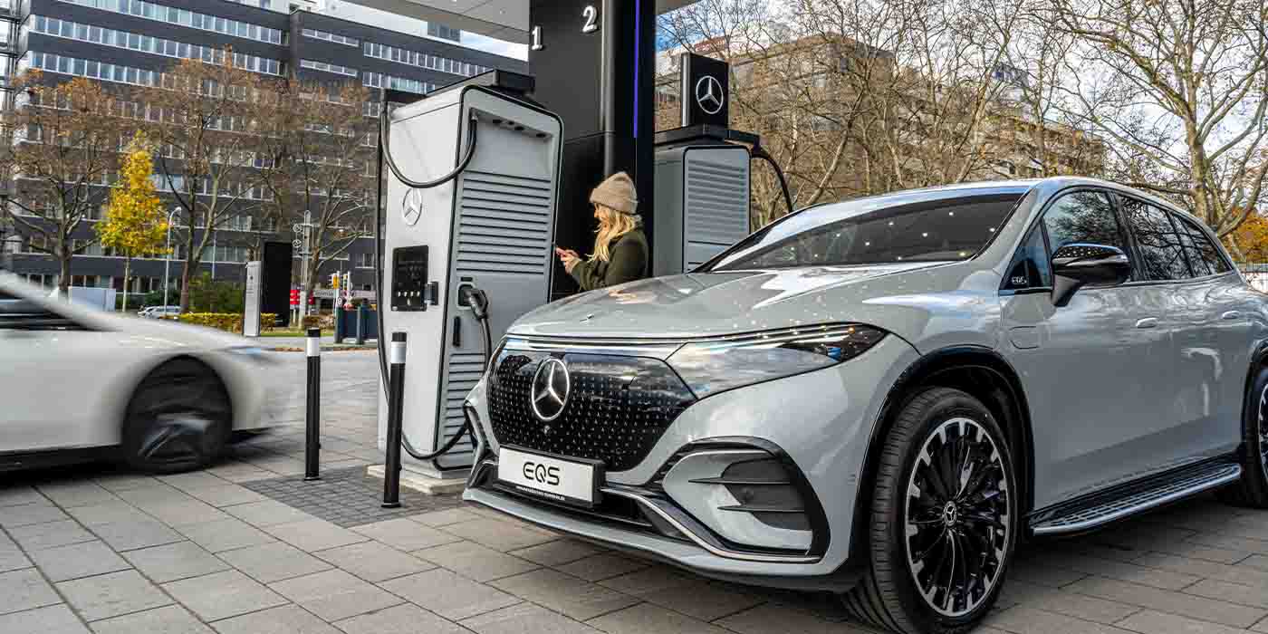 Une Mercedes-Benz électrique de couleur claire en train de charger à une borne de recharge publique dans une rue européenne. L'architecture en arrière-plan est typiquement européenne.