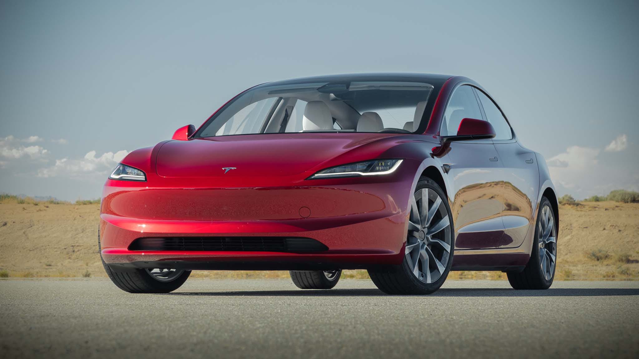 Vue de face d'une Tesla Model 3 Highland 2026 de couleur sombre, garée dans une rue de ville au crépuscule, ses phares allumés.