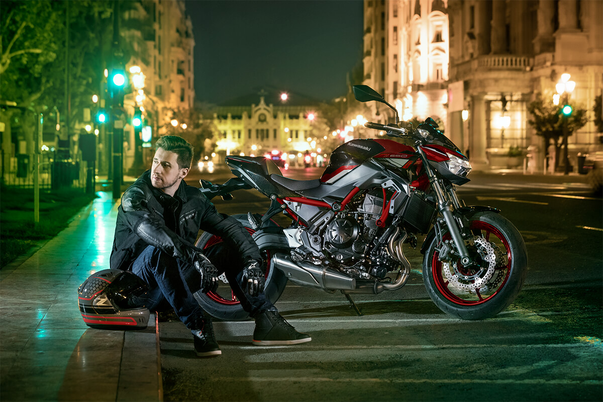 Une Kawasaki Z650 rouge et noire circulant dans une rue de ville animée, avec des bâtiments modernes en arrière-plan.