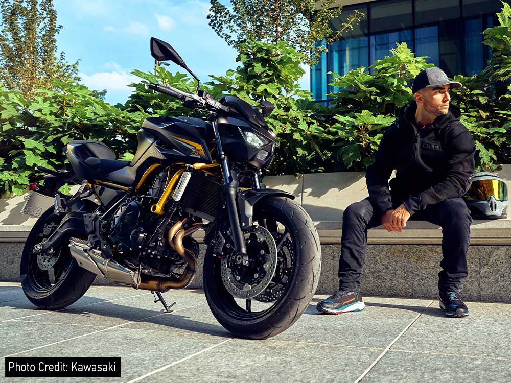 Une Kawasaki Z650 verte et noire en pleine accélération sur une route de campagne sinueuse, avec des arbres et des collines en arrière-plan.