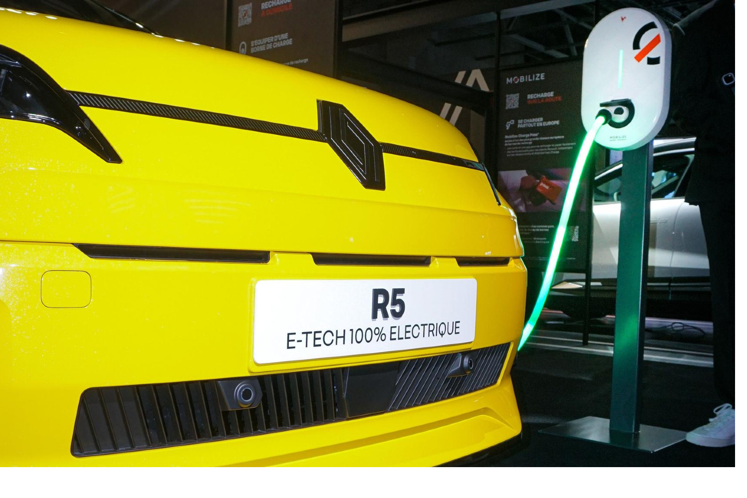 Une Renault 5 électrique blanche est branchée à une borne de recharge rapide sur un parking urbain, avec des bâtiments modernes en arrière-plan.