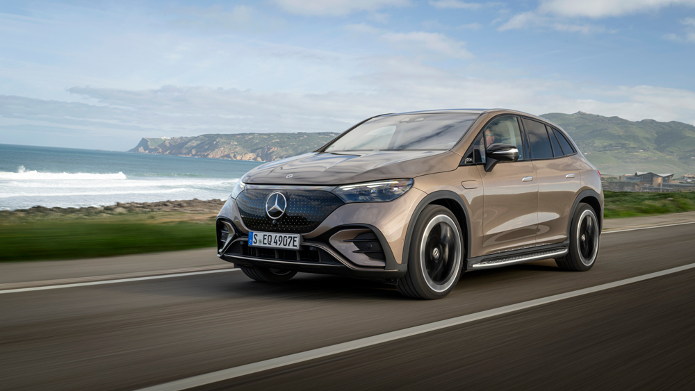 Un SUV Mercedes-Benz EQE gris foncé roule sur une route de campagne sinueuse, entouré de verdure luxuriante et de montagnes lointaines sous un ciel bleu clair.