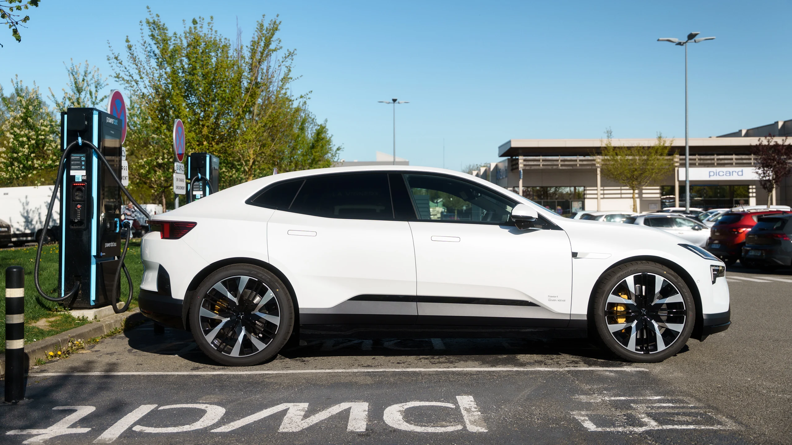 Une voiture électrique blanche est branchée à une borne de recharge Powerdot dans un environnement urbain, avec le logo Octopus Energy visible sur la borne.