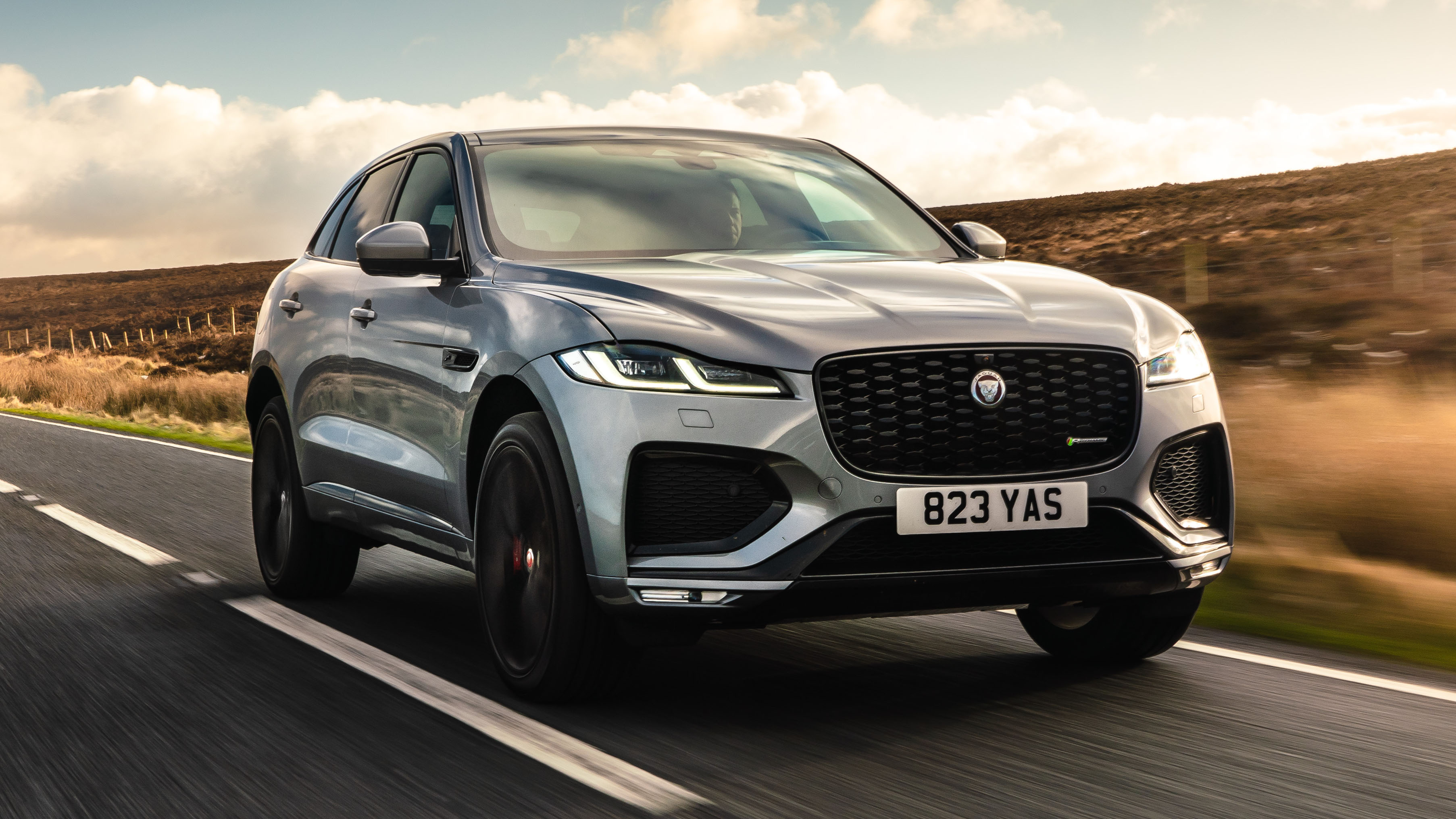 Un SUV Jaguar F-Pace de couleur sombre, garé sur une route de campagne, avec des arbres en arrière-plan. Le véhicule est élégant et moderne.
